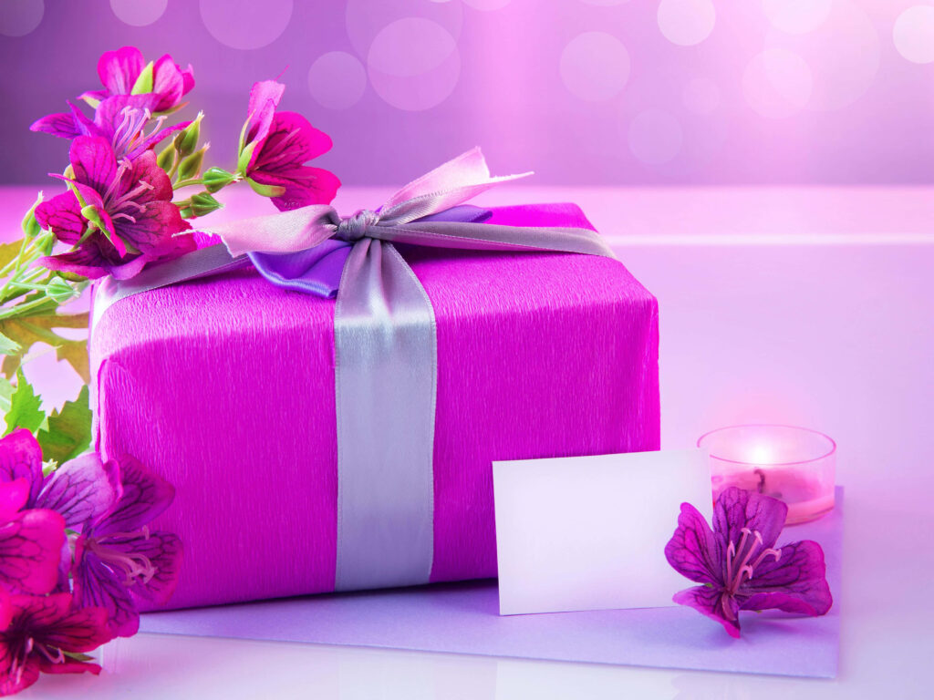 Purple gift box with silver ribbon surrounded by flowers candle and blank card on pink background Wallpaper