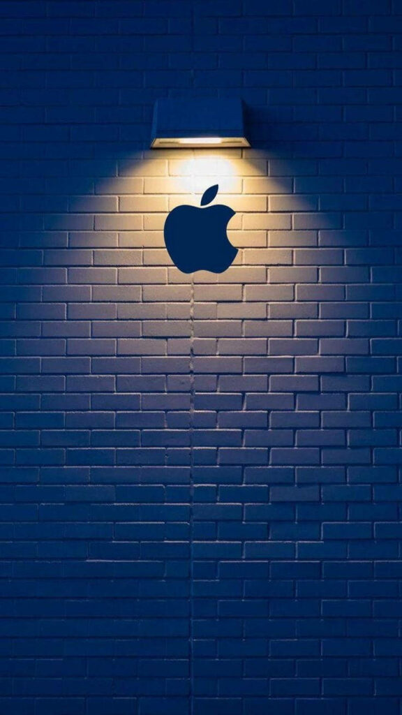Golden Glow: Stunning Black Apple Logo iPhone Wallpaper Illuminated by a Yellow Streetlight against a Brick Wall Background