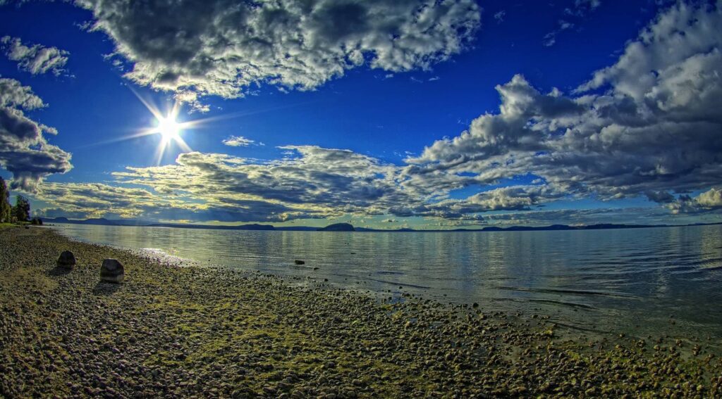 Radiant sun over tranquil coast with smooth stones dramatic clouds and vibrant blue sky wallpaper