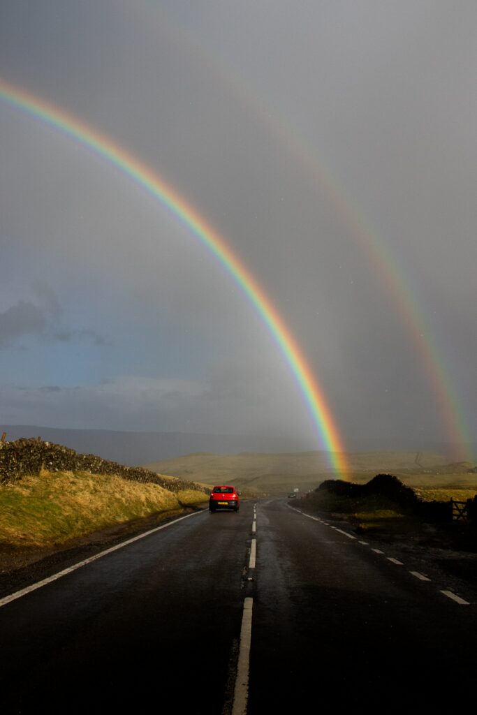 Rainbow over cloudy sky and wet country road with red car driving near London landscape Wallpaper