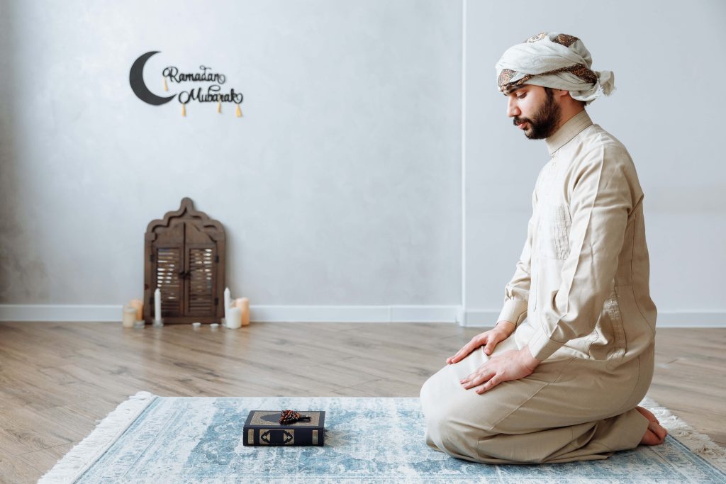 Bearded man in beige thobe praying on rug with Quran and beads Ramadan Mubarak wallpaper background