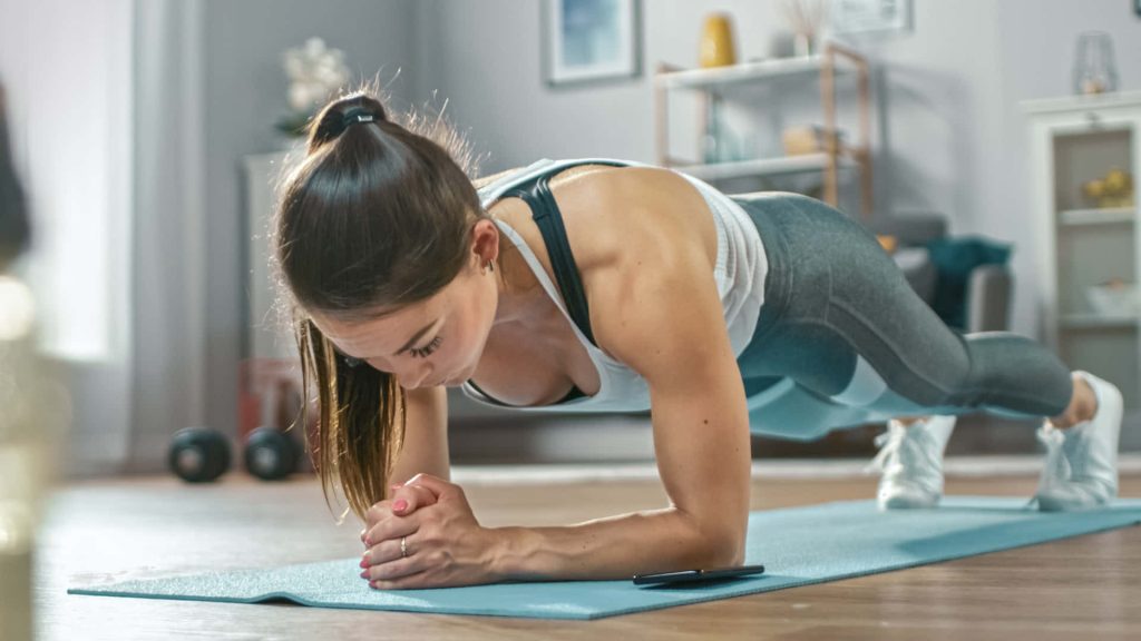 Woman doing forearm plank on blue yoga mat in bright modern living room fitness wallpaper