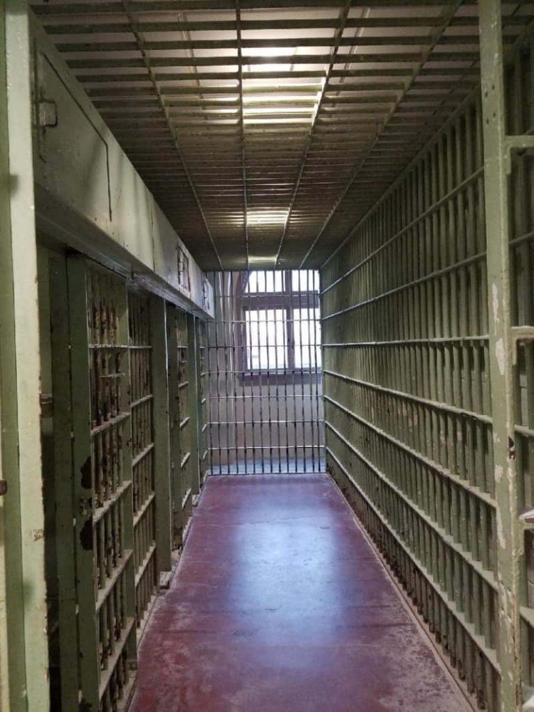 Realistic prison cell block hallway with metal bars dark red floor and natural light window Wallpaper