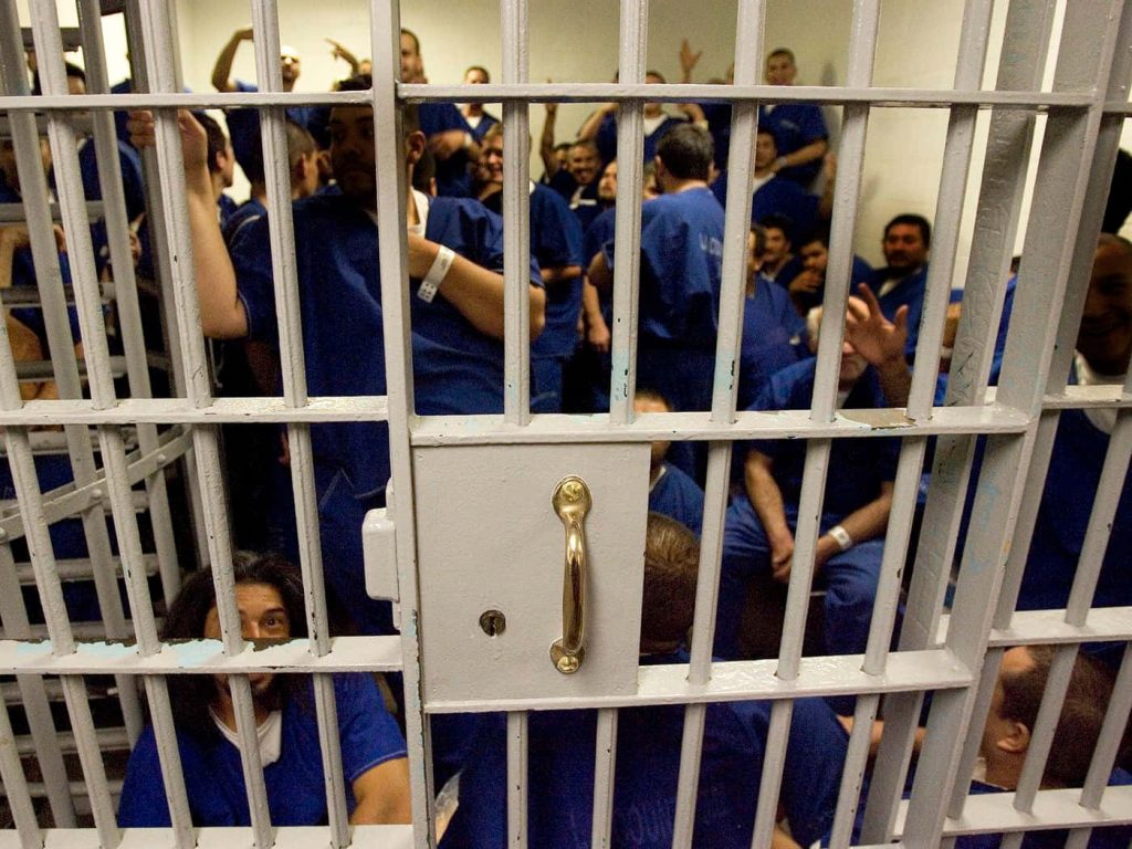 Group of inmates in blue uniforms behind metal jail bars prison cell background wallpaper