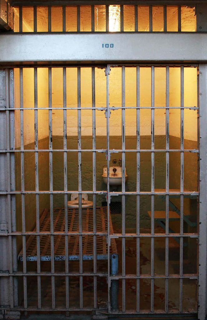 Realistic prison cell wallpaper with barred sliding doors cot washbasin and dim lighting background