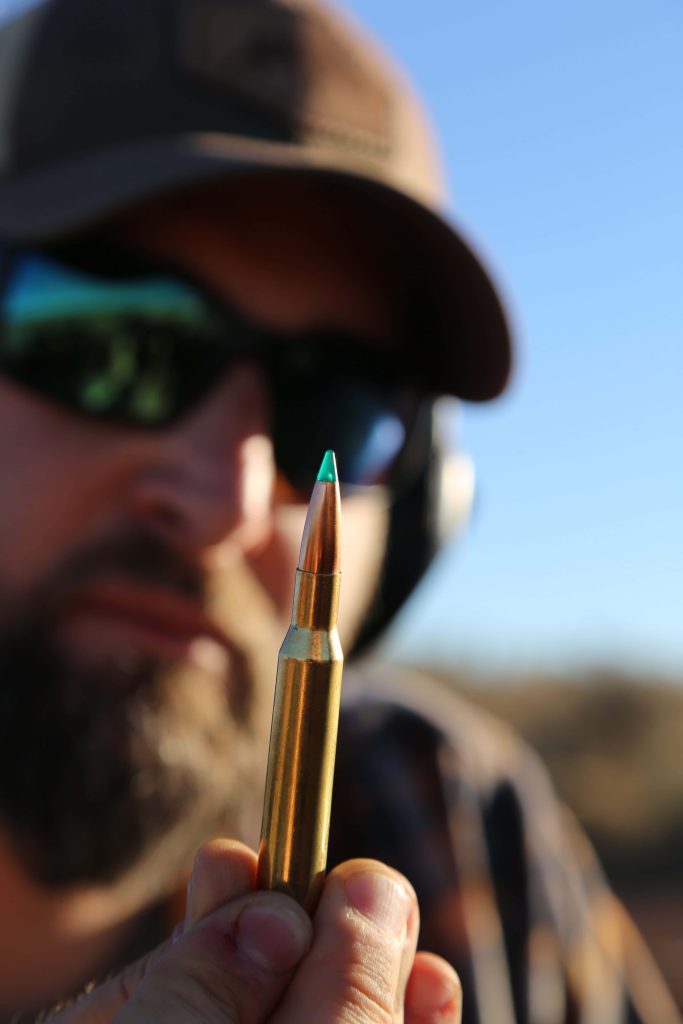Close-up of rifle cartridge with green polymer tip and blurred bearded man in background wallpaper