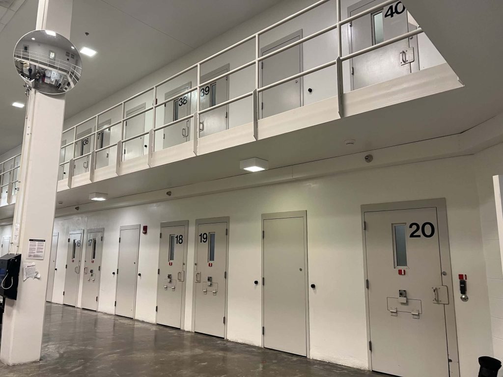 Two-tier prison corridor with white walls numbered metal cell doors security mirror bright lighting Wallpaper