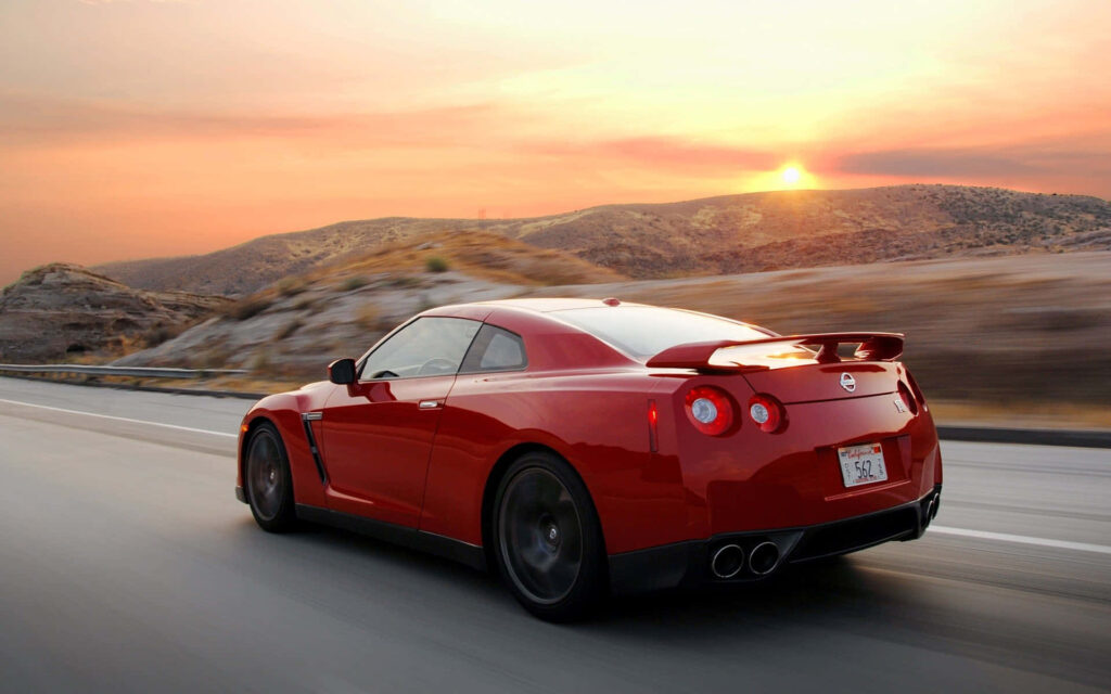 Red 2009 Nissan R35 GTR speeding on open road at sunset with rugged hills and warm sky background Wallpaper
