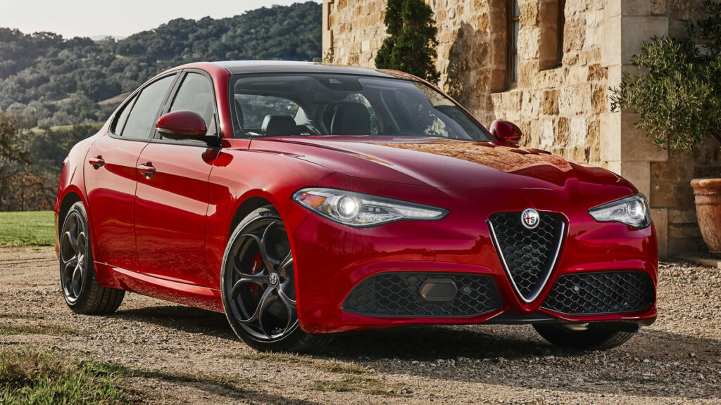 Red Alfa Romeo Giulia sports car on scenic mountain road with greenery and stone architecture Wallpaper