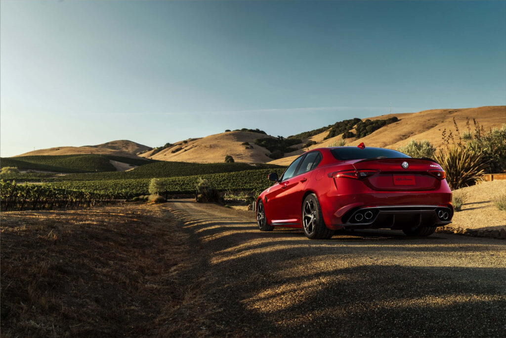 Red Alfa Romeo Giulia speeding on winding countryside road with golden hills and clear sky Wallpaper