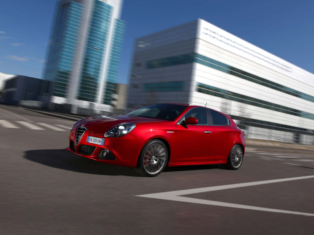 Red Alfa Romeo Giulietta speeding in modern cityscape with sleek design and glass buildings background Wallpaper