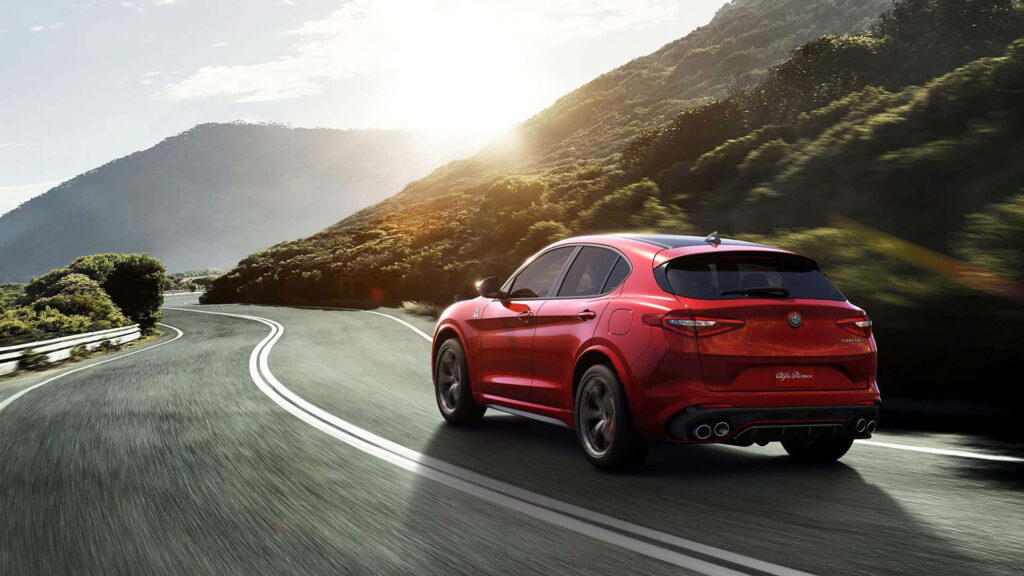 Red Alfa Romeo Stelvio driving on scenic road with green hills and sunny mountain background Wallpaper