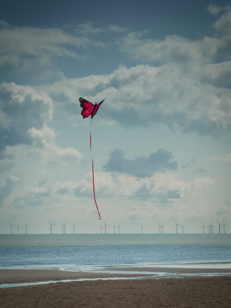 Red butterfly kite flying over calm beach with sea wind turbines and partly cloudy sky background Wallpaper