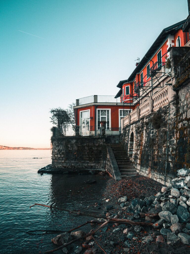 Red coastal building with stone walls on rocky shore driftwood calm sea and blue sky wallpaper