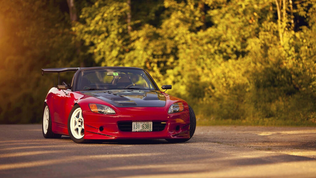 Red Honda S2000 with sporty body kit and large wing parked on sunlit road with green trees Wallpaper