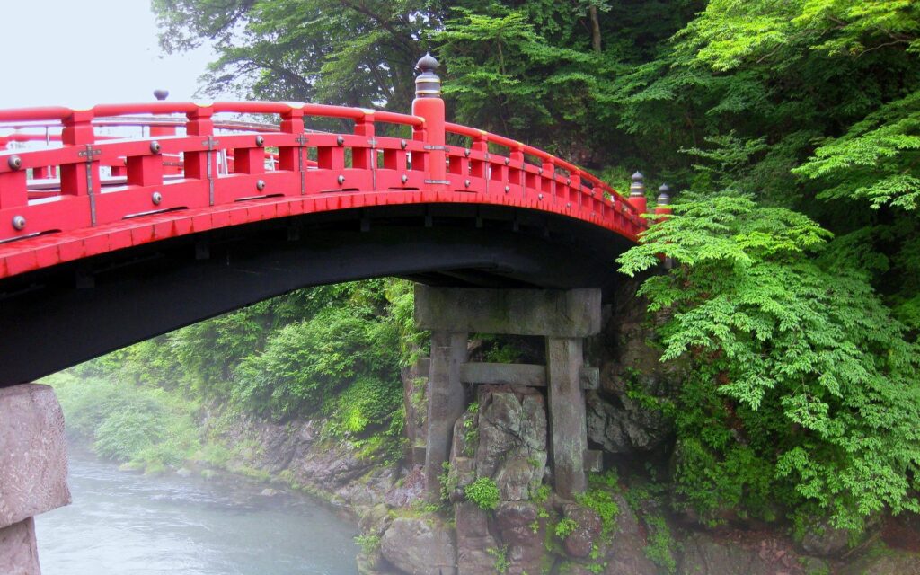 Traditional red Japanese bridge over calm river surrounded by lush green trees wallpaper background