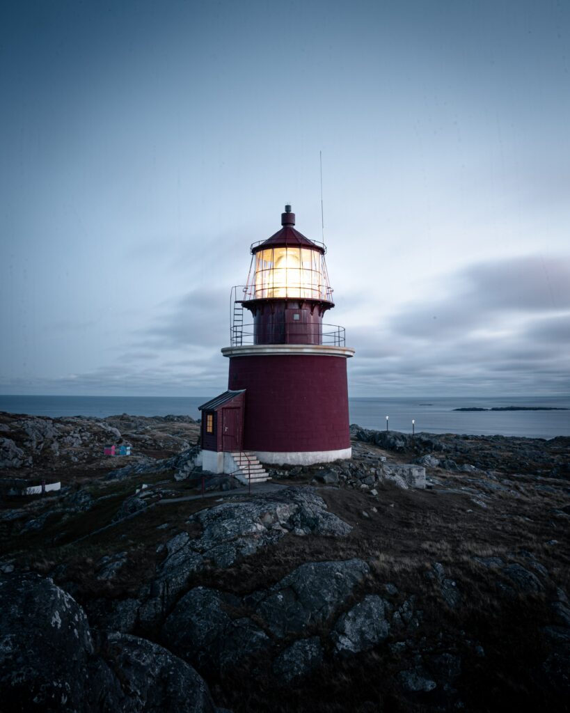 Red lighthouse glowing at dusk on rugged coastal rocks with vast sea and moody sky background Wallpaper