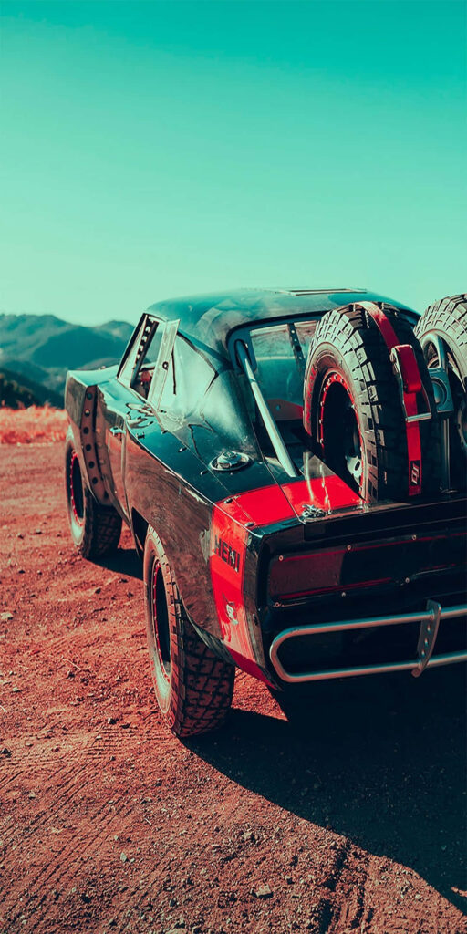 Red muscle car with off-road tires and spare wheels on rugged terrain under blue sky wallpaper