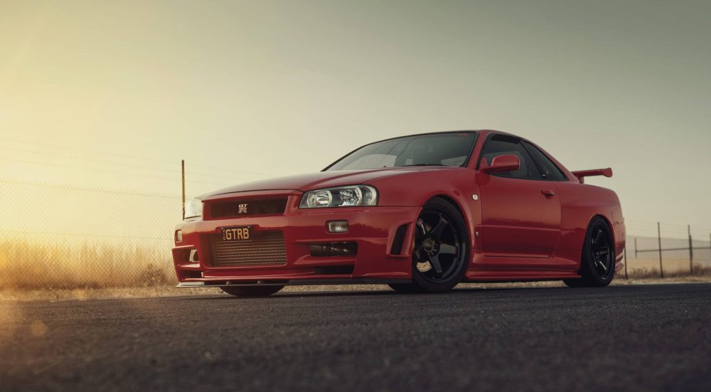 Red Nissan Skyline GT-R R34 at sunset on open road with sleek black rims wallpaper background