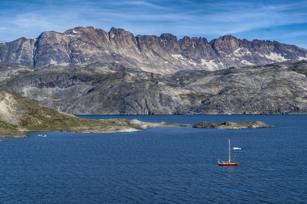 Red sailboat sailing on deep blue Greenland Sea with dramatic gray mountains and clear sky background Wallpaper