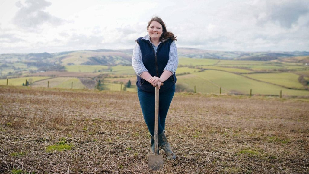 Farmer Hannah Jackson the Red Shepherdess in practical gear posing with shovel in rural fields Wallpaper