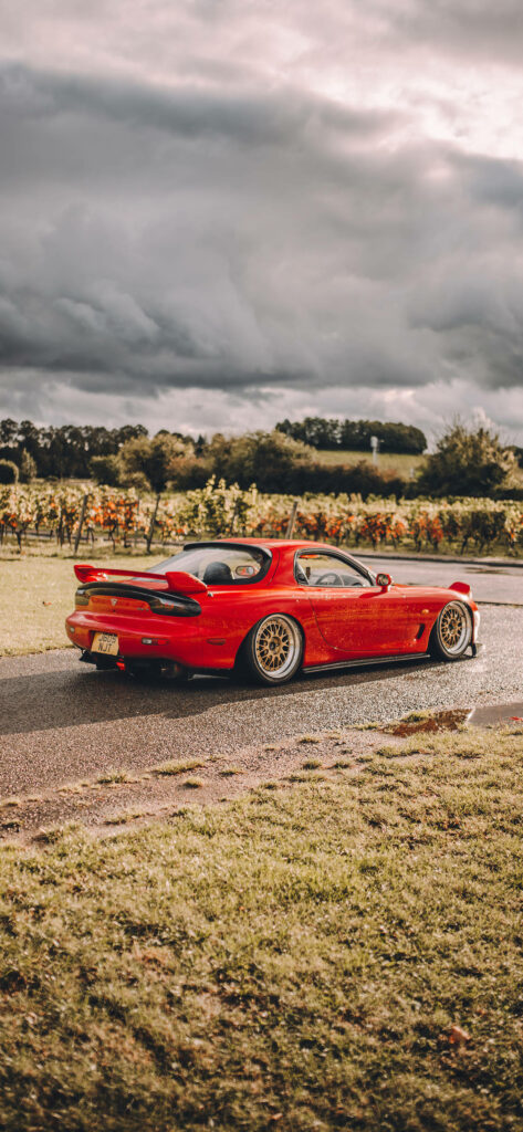 Red sports car driving fast on scenic country road with green fields and cloudy sky background Wallpaper