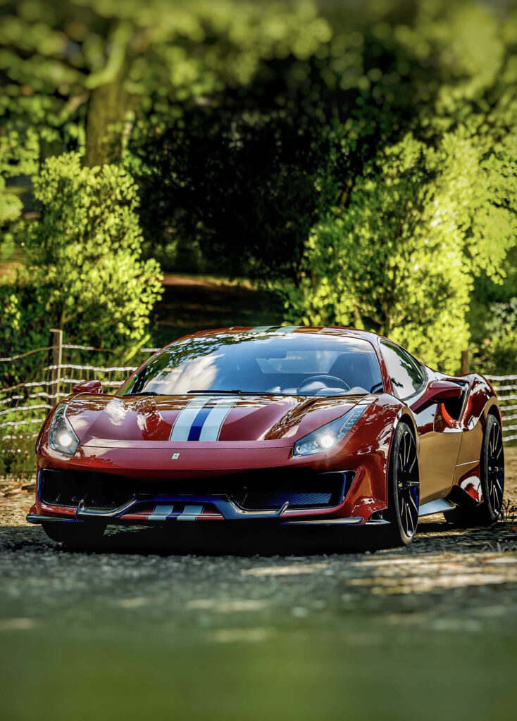 Red sports car with blue white racing stripe parked on sunny path surrounded by green trees Wallpaper