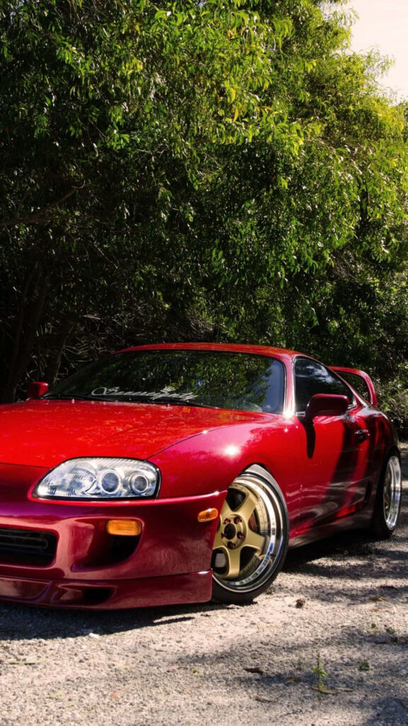 Red sports car with golden rims on sunlit road surrounded by vibrant green trees wallpaper