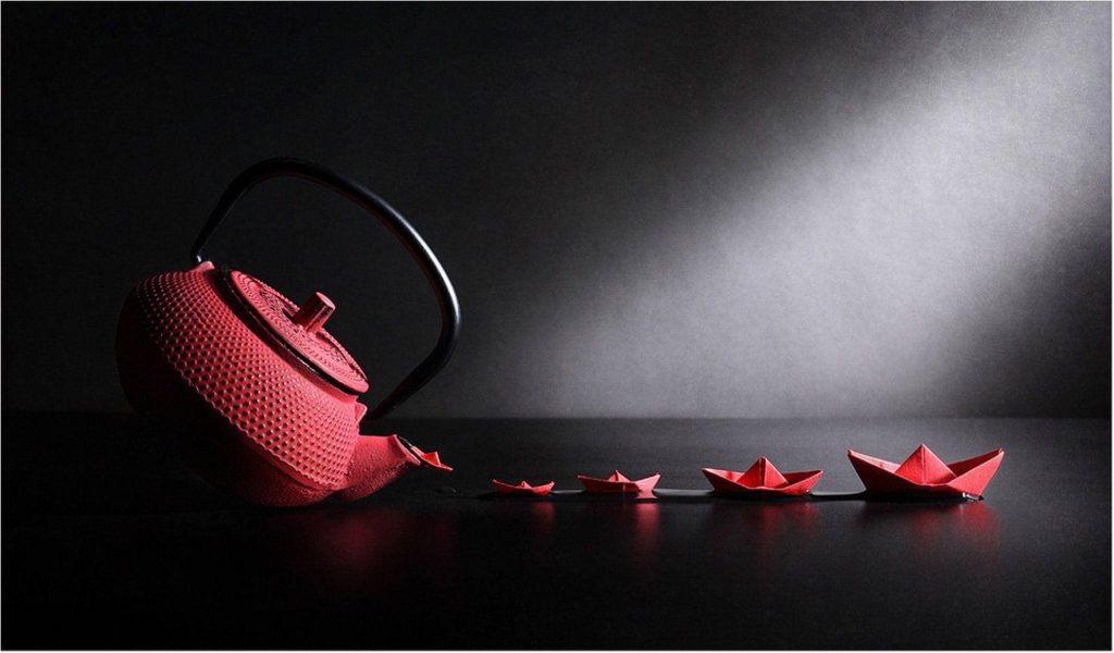 Red textured teapot pouring dark liquid with floating red paper boats on dark background wallpaper