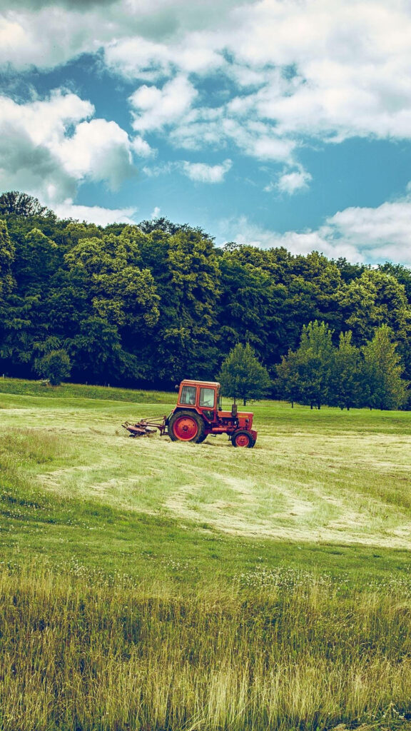 Red tractor with flail mower in freshly mowed field surrounded by green trees under cloudy sky Wallpaper
