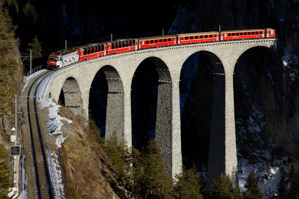 Red train crossing tall arched stone railway bridge over forested hills and rugged cliffs landscape Wallpaper