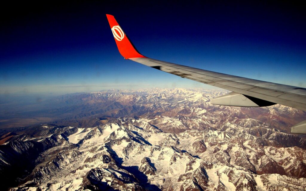 Airplane wing with red tip above snow-capped mountains under deep blue sky travel wallpaper background
