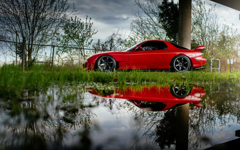 Roaring Red: An Rx-7's Reflection Captured on a Water Mirage, Surrounded by Nature's Canopy and Metal Perimeter Wallpaper