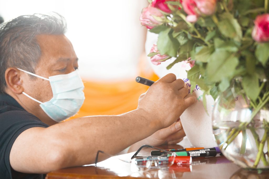 Man in surgical mask writing on memorial urn with pink roses and glasses funeral background Wallpaper