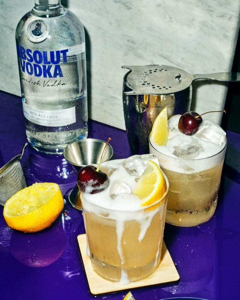 Absolut Vodka lemon cocktail with ice cherries and tools on vibrant purple background wallpaper