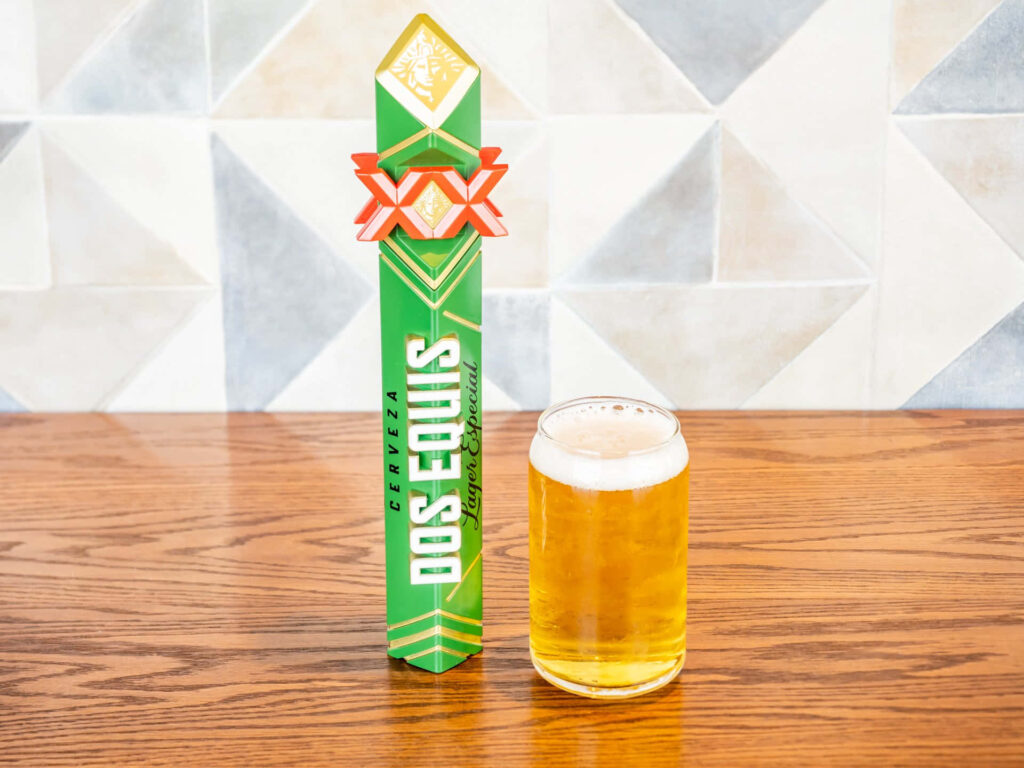 Dos Equis beer tap next to a golden poured glass on wooden table with modern tiled backdrop Wallpaper