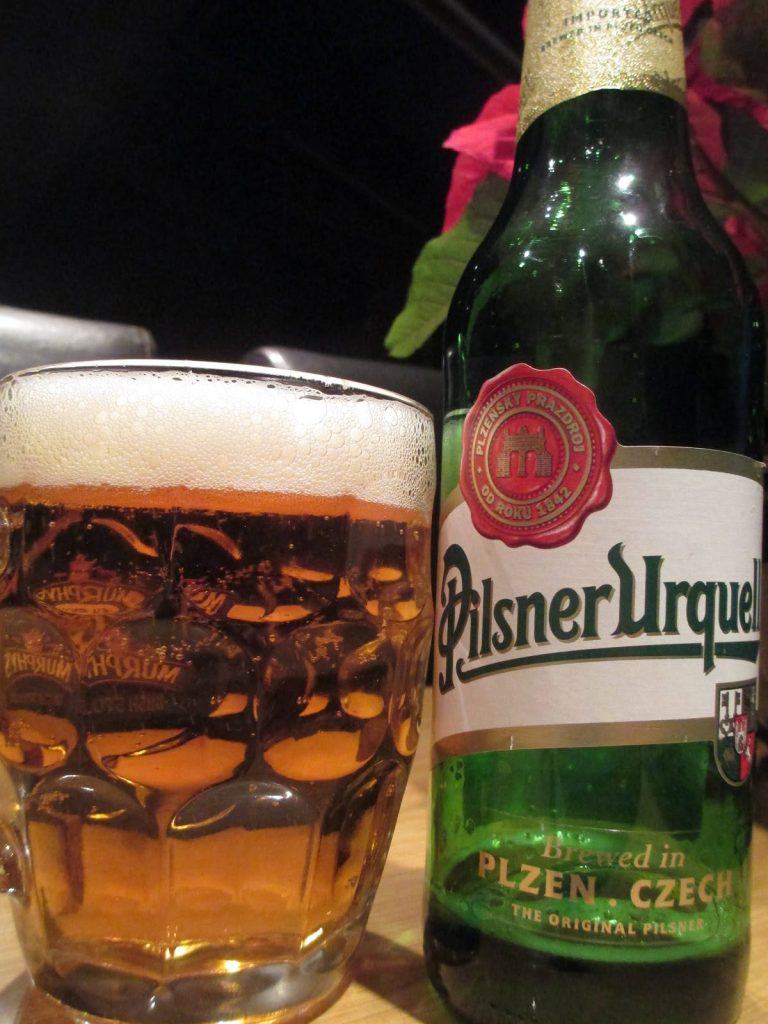 Golden Pilsner Urquell beer in dimpled pint glass with green bottle and frothy head close-up Wallpaper
