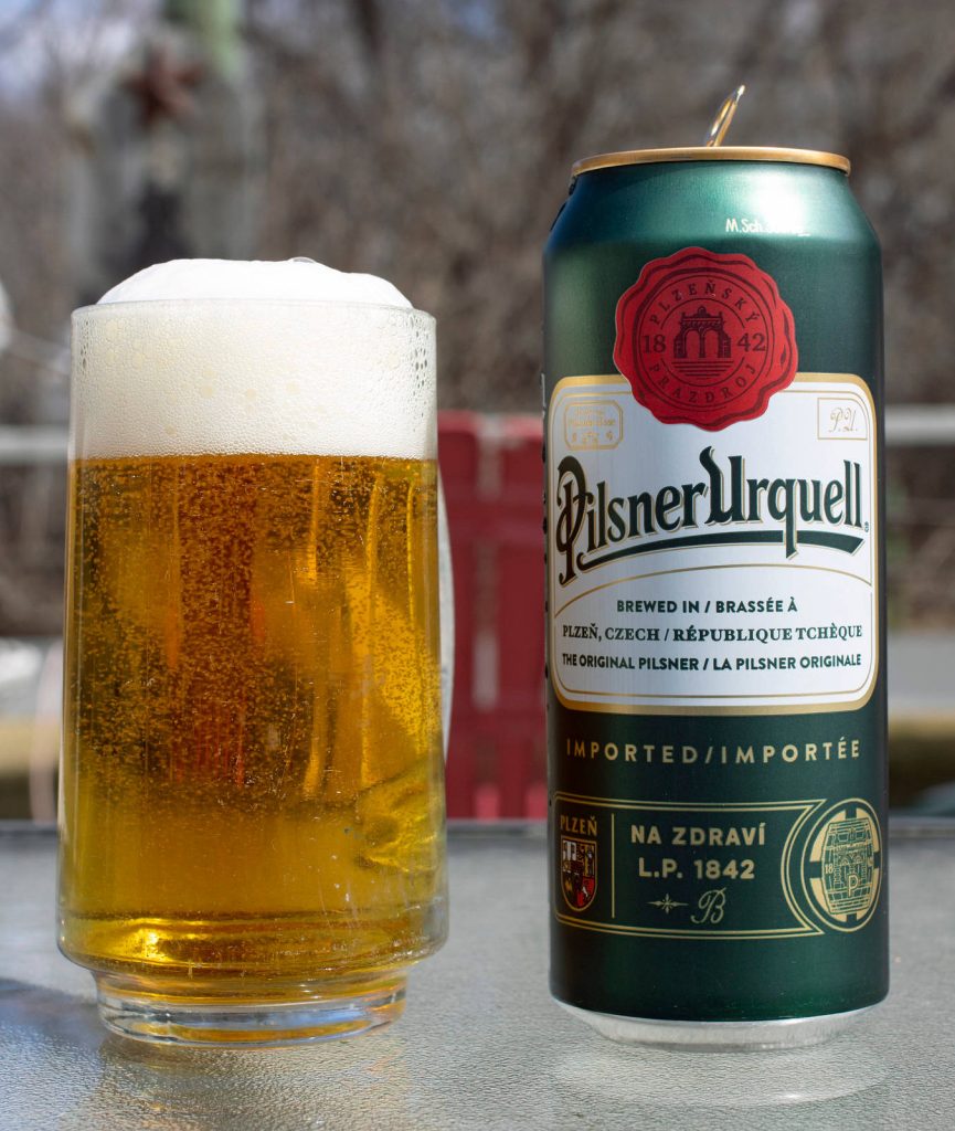 Glass of frothy Pilsner Urquell lager beside green can on glass table in bright outdoor setting Wallpaper