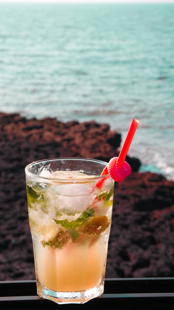 Refreshing tropical cocktail with mint lemon ice cubes red straw ocean rocky shore background Wallpaper