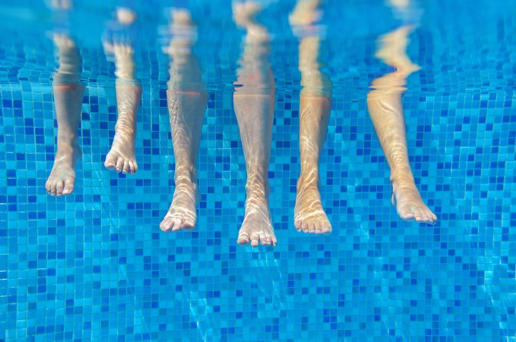 Three pairs of legs dip into clear water over blue mosaic pool tiles summer swimming scene Wallpaper