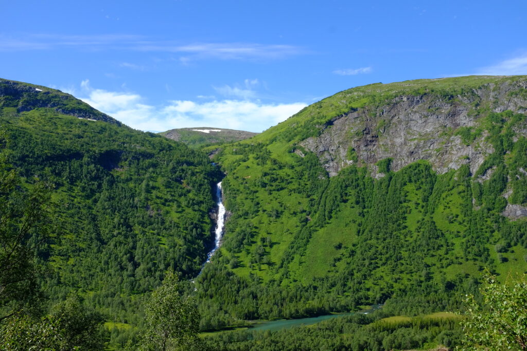 Waterfall cascading down green mountainside with dense forest and bright blue sky background Wallpaper