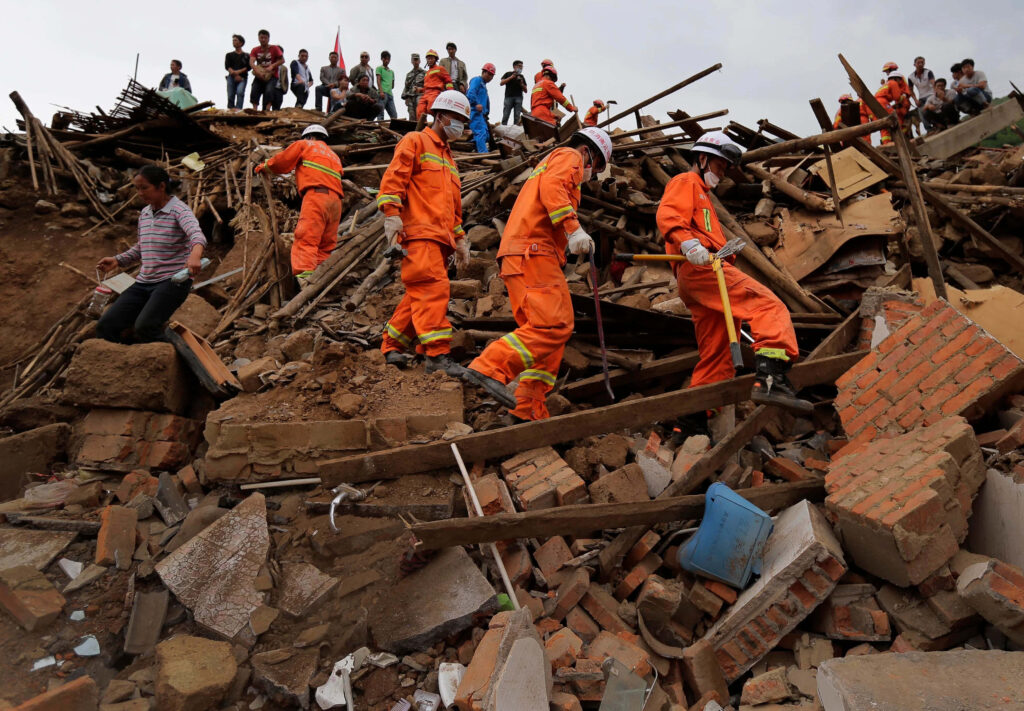 Rescue workers in orange uniforms search earthquake rubble with onlookers above broken bricks Wallpaper