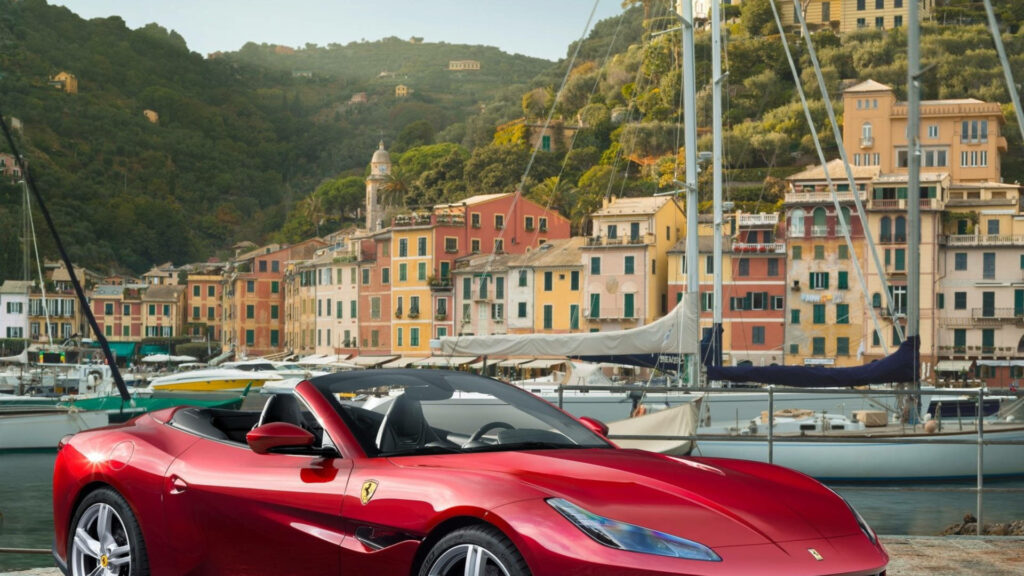 Speed and Vibrance: A Fiery Ferrari Accelerates Near a Vibrant Portside Community Wallpaper