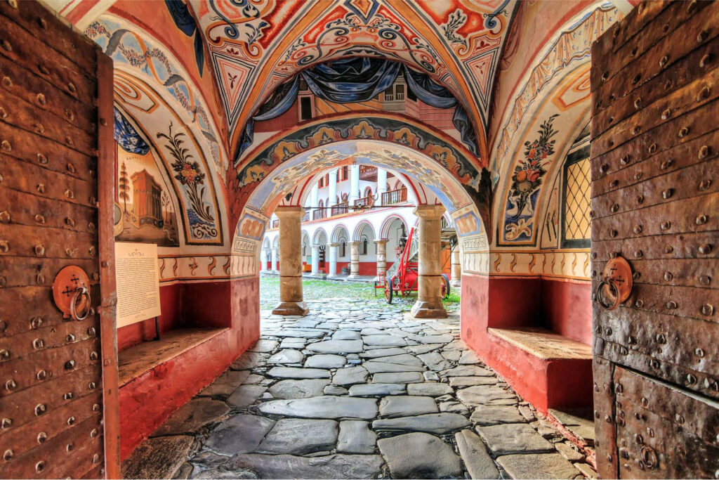 Rila Monastery hallway with vibrant frescoes and ornate archway overlooking historic courtyard wallpaper