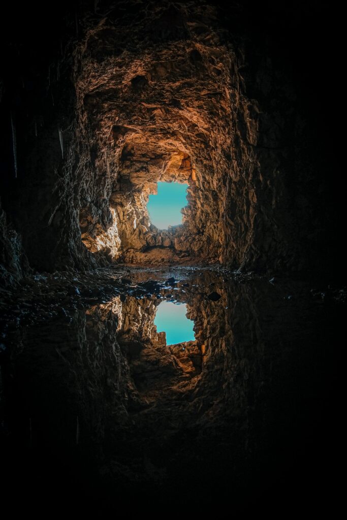Rocky cave tunnel with bright sky reflection in puddle highlighting textured walls depth Wallpaper
