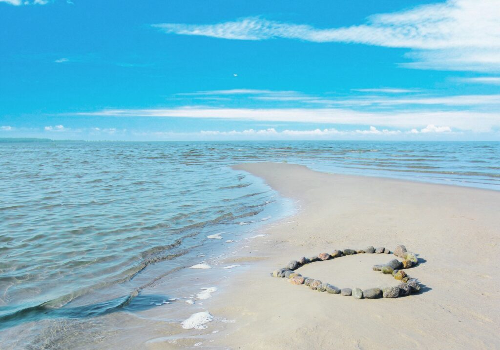 Heart shape of smooth rocks on serene beach with gentle waves and vibrant blue sky background Wallpaper