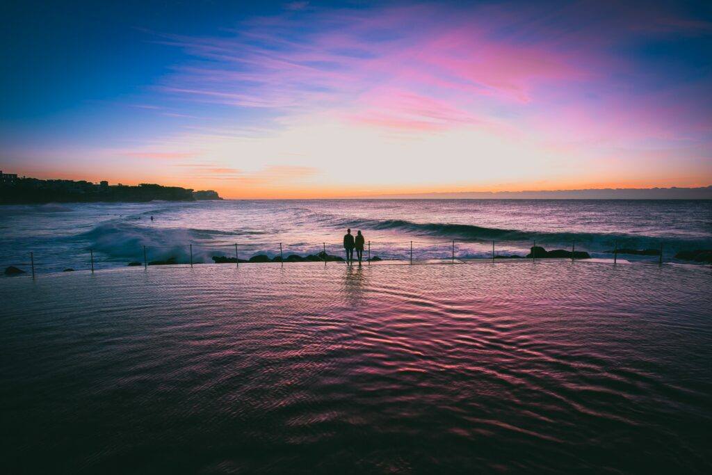 Romantic couple at infinity pool edge viewing vibrant pink purple blue sunset over calm sea Wallpaper