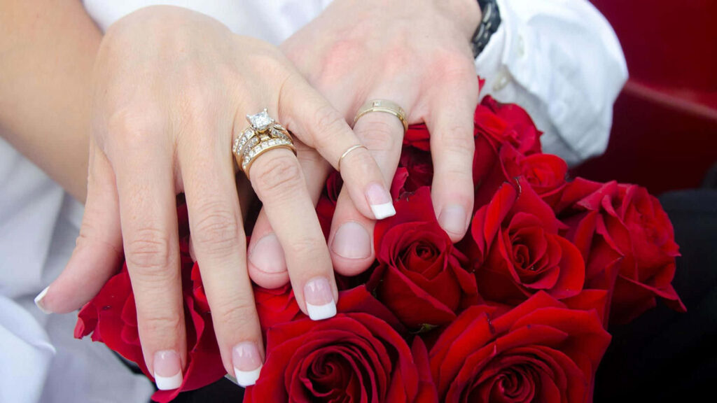 Romantic couple hands with elegant engagement rings over vibrant red rose bouquet wallpaper background