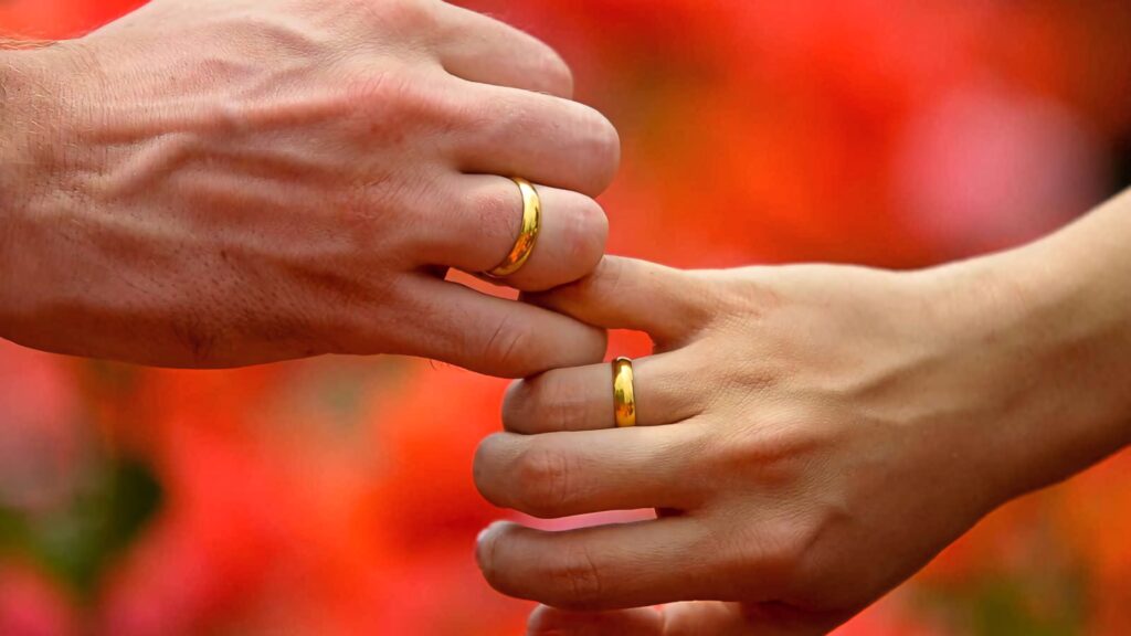 Two hands with gold band engagement rings on vibrant red and orange floral romantic wallpaper