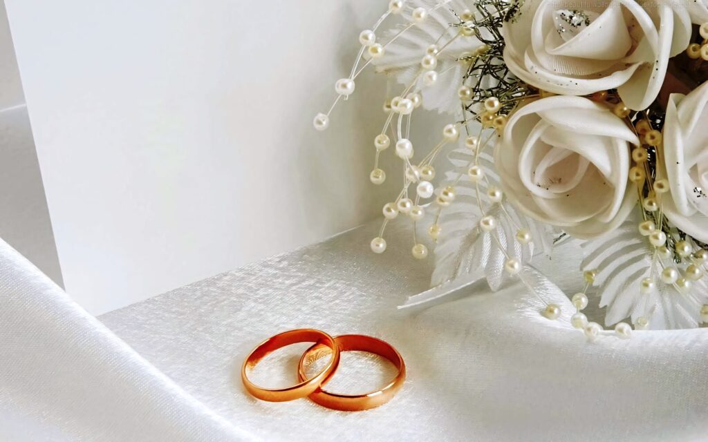 Two gold engagement rings on white satin with white roses and pearls romantic wedding background Wallpaper
